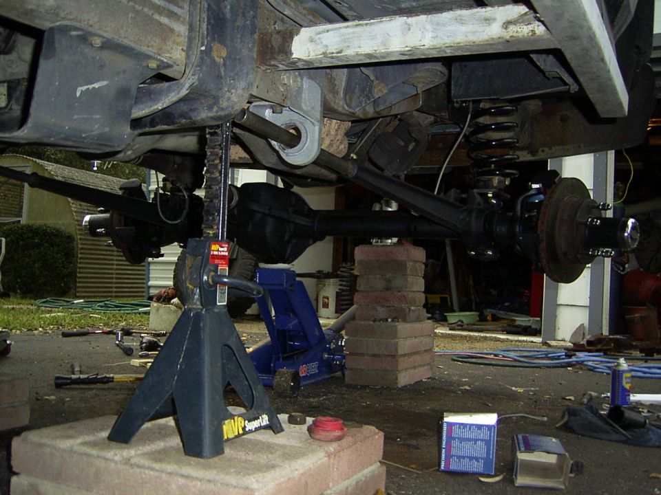 Vehicle undercarriage with suspension and axles supported by jack stands and bricks. Tools and parts are scattered around
