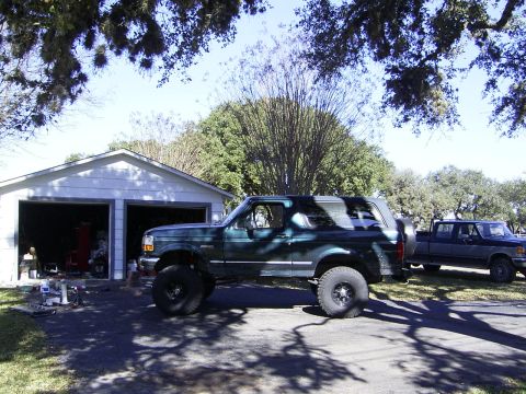 1995 Ford Bronco - A lifted 1995 Ford Bronco sits in front of a garage, with a pickup truck parked nearby.