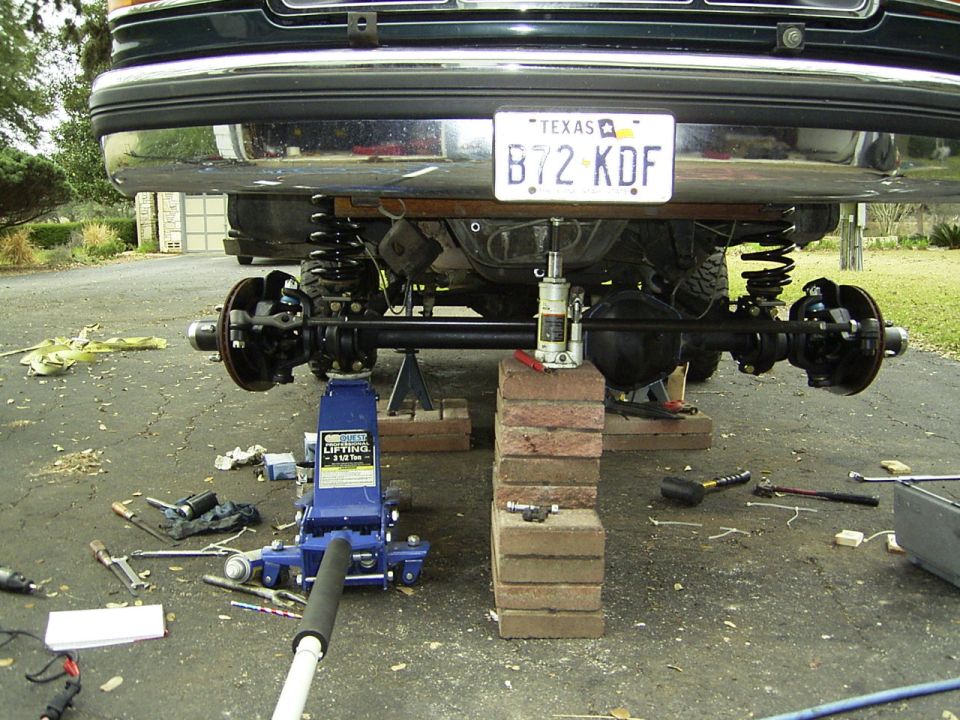 Rear axle and suspension of a vehicle being worked on, supported by jacks and bricks. Texas license plate B72 KDF is visible