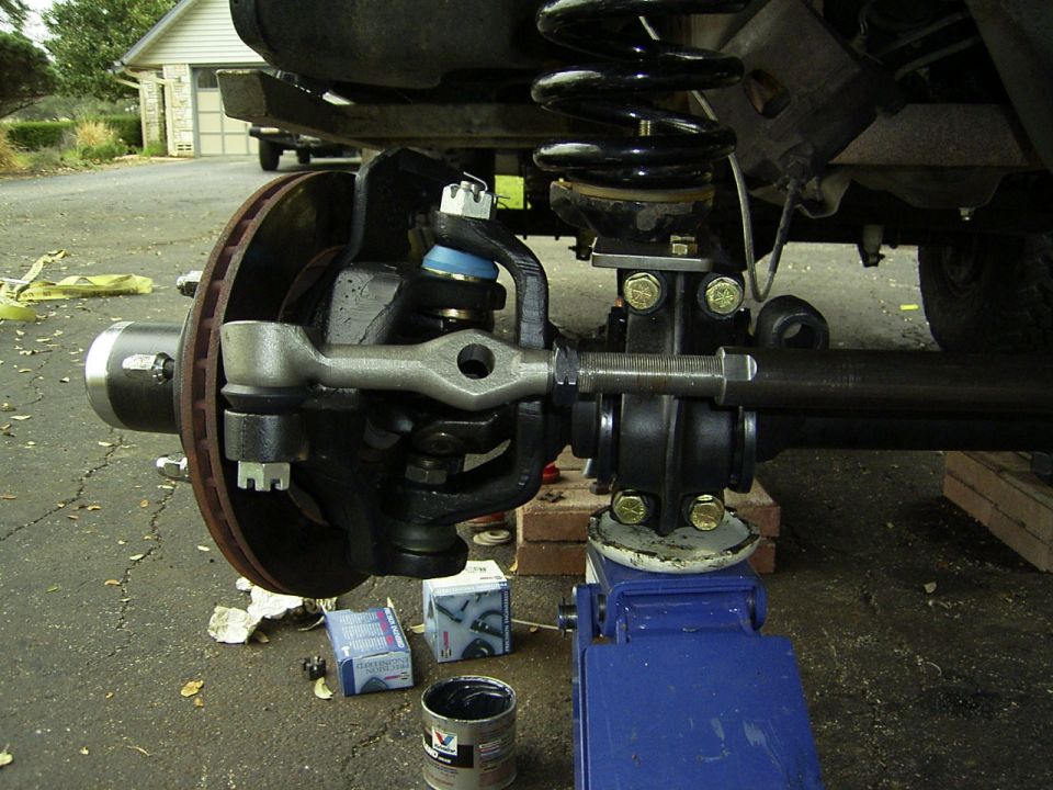 Close-up of a vehicle's front suspension and steering linkage, with a blue jack supporting the frame