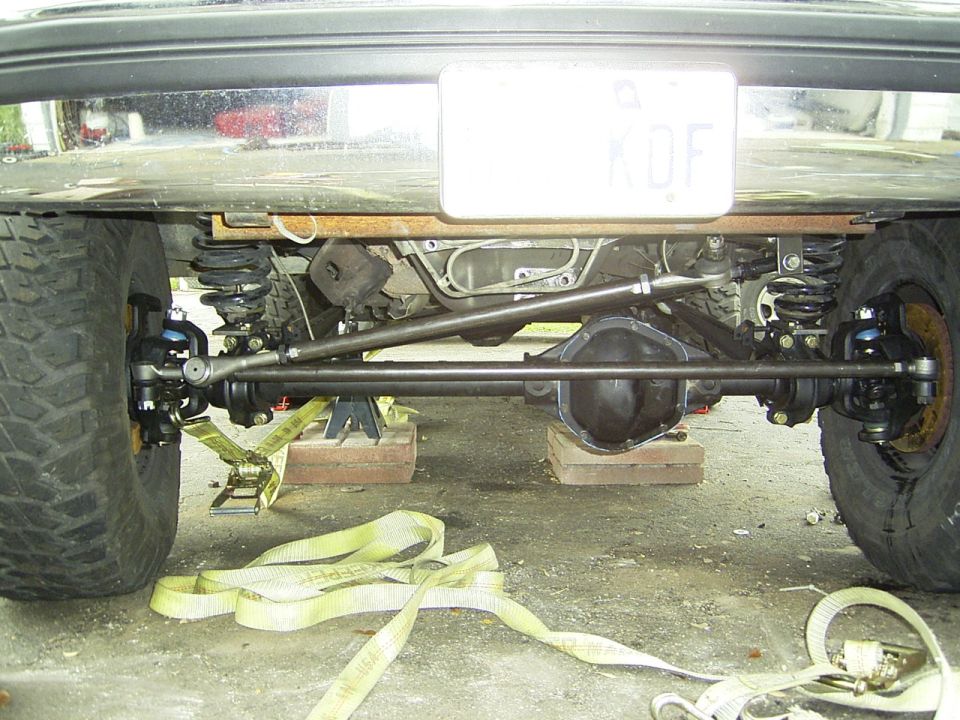 Underside view of a vehicle's rear axle, suspension, and steering components, supported by jack stands