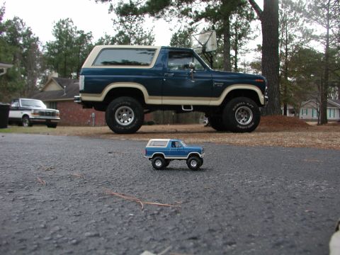 1985 Ford Bronco - A full-size blue Bronco is parked next to a miniature toy Bronco, with another truck in the background.