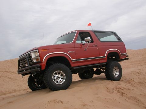 1984 Ford Bronco - A red Ford Bronco with a black roof and trim is shown lifted on large tires, navigating sand dunes under a cloudy sky.