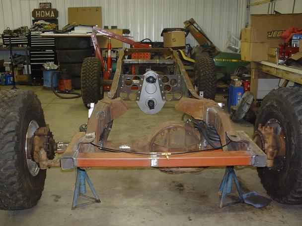 Rusty truck frame on jack stands with front axle, orange crossmember and fabricated dash panel in a garage