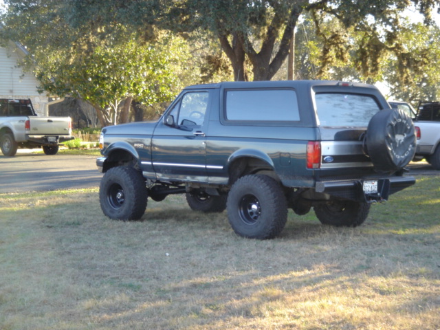 Dark green lifted Ford Bronco parked on grass, another truck in background