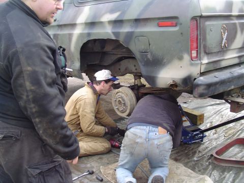 1990 Ford Bronco - Three individuals attend to the rear suspension of a lifted 1990 Ford Bronco, which is painted in camouflage.