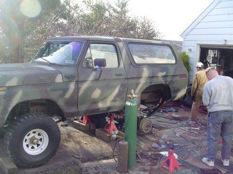 1990 Ford Bronco - Ford Bronco undergoing repairs, lifted on jacks with rear axle removed, in a cluttered outdoor workshop.