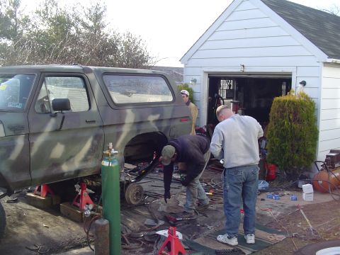 1990 Ford Bronco - Men work on a camouflaged Ford Bronco lifted on jack stands in front of a garage.