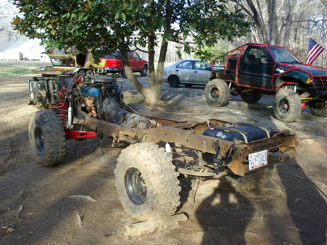 Bare vehicle frame with mud tires and exposed engine in dirt yard, other vehicles behind