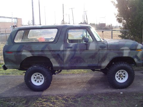 1990 Ford Bronco - Camouflaged Ford Bronco sits on a paved surface with a chain-link fence in the background.
