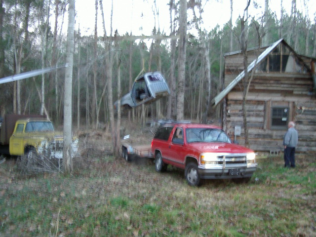 Red Chevy SUV with flatbed trailer hauling a vehicle in woods, old log cabin in background
