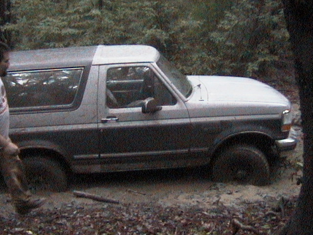 Muddy two-tone Ford Bronco stuck to its rockers in a wooded mud hole, side view