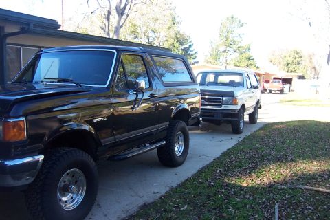 1989 Ford Bronco - Two Ford Broncos, a black one and a white one, are parked side-by-side on a paved driveway.