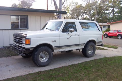 1990 Ford Bronco - A white Ford Bronco is parked in a driveway next to a house, with a red pickup truck visible in the background.
