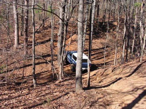 1978 Ford Bronco - A blue and white SUV navigates a wooded trail, its white camper shell visible.