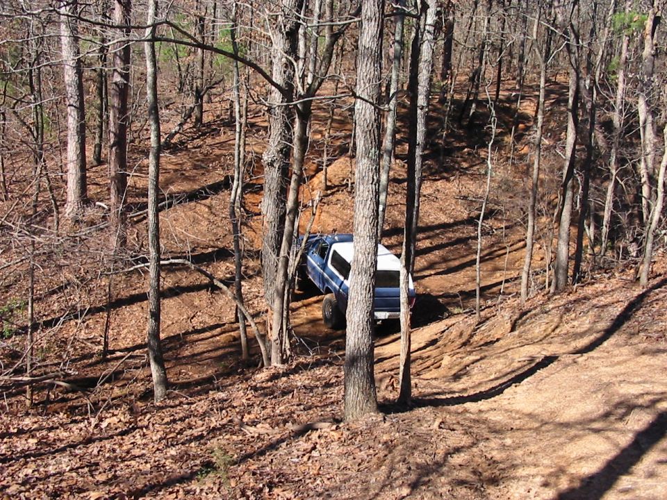 Blue and white SUV with a white camper shell driving off-road on a dirt path in a wooded area