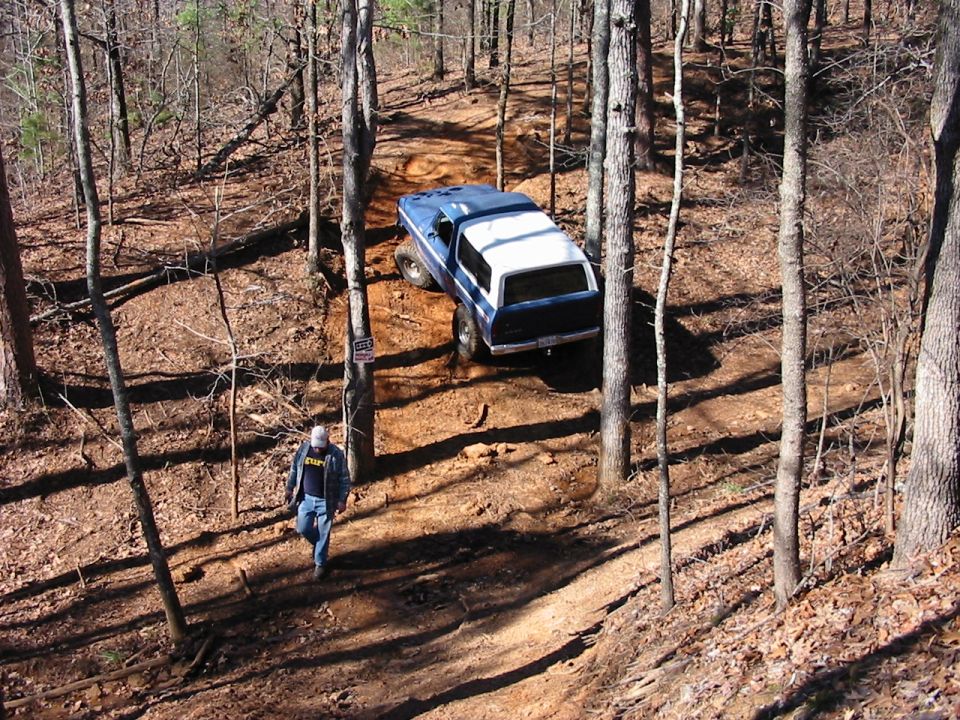 Blue and white SUV driving on a dirt trail in a wooded area. A person walks on the trail below the vehicle