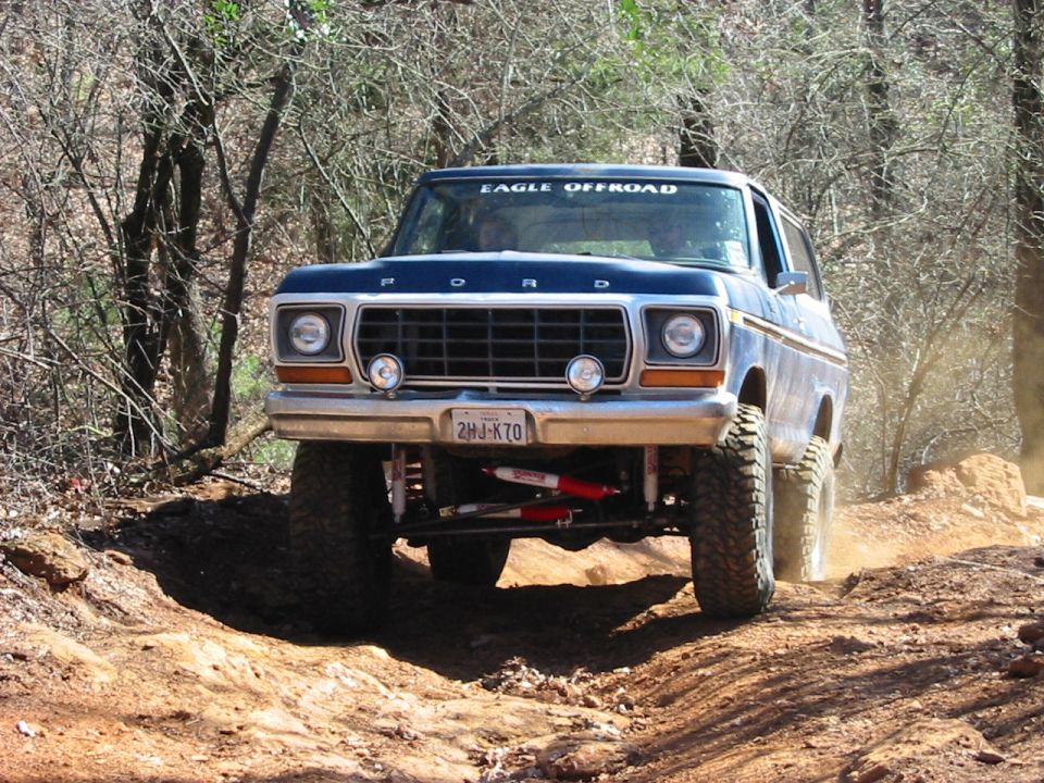 Blue 1978 Ford Bronco with lifted suspension and off-road tires driving on a dirt trail
