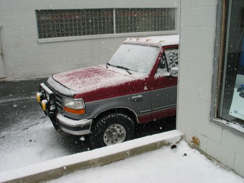 1995 Ford Bronco - A two-tone red and gray pickup truck is partially obscured by snow and a building corner.