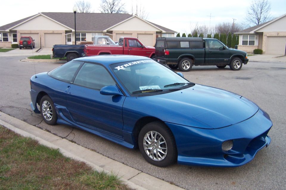 Blue Ford Probe coupe with body kit parked at a curb, trucks and houses behind