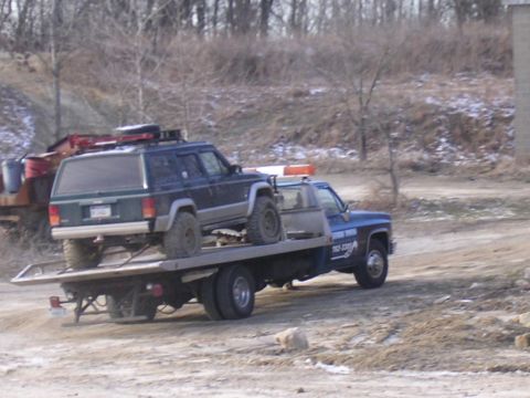 1995 Jeep Cherokee - A tow truck transports a Jeep Cherokee on its flatbed, with a wooded area in the background.