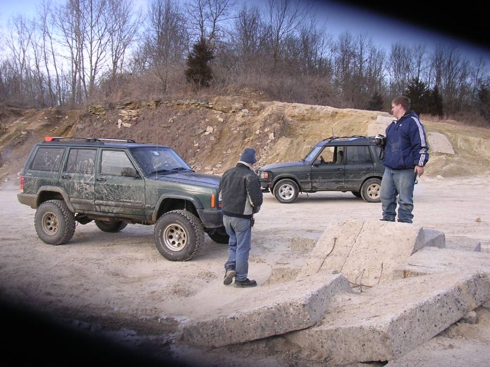 Green Jeep Cherokee and a dark green SUV parked on dirt with two men standing nearby