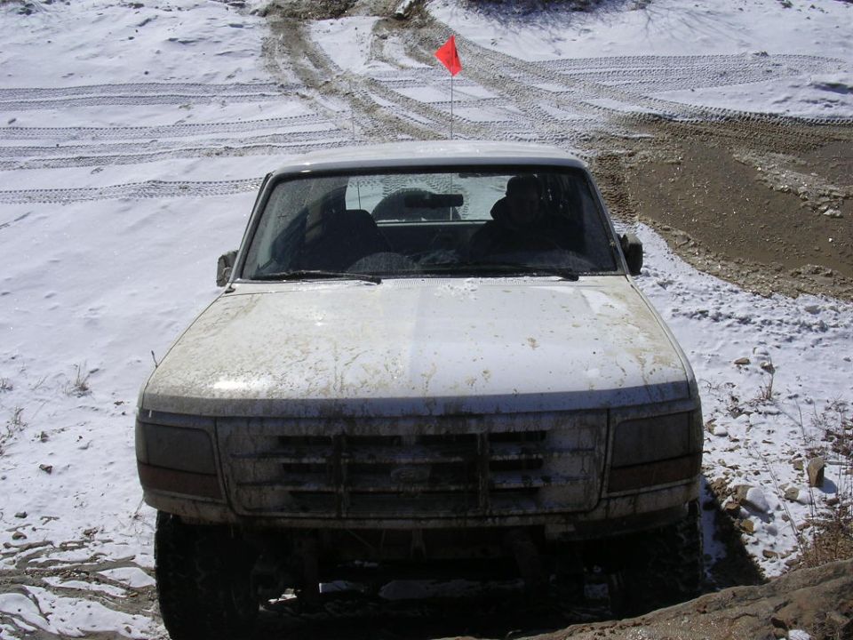 White pickup truck with a red flag on top, parked on a snowy, muddy trail. Front view
