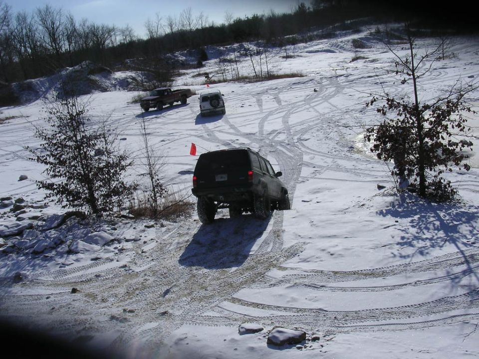 Black SUV on snowy hill, rear view. Other trucks in background. Tire tracks visible
