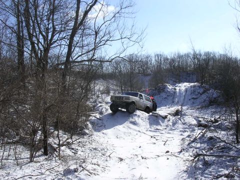 1995 Jeep Cherokee - A white SUV navigates a challenging snowy terrain, with bare trees and a clear blue sky overhead.
