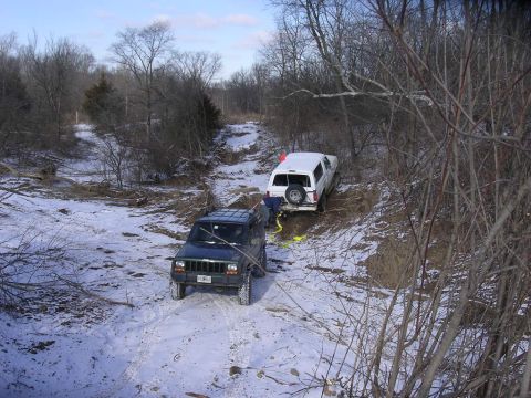 1995 Jeep Cherokee - Two off-road vehicles navigate a snowy, wooded trail, with one truck appearing to be stuck.