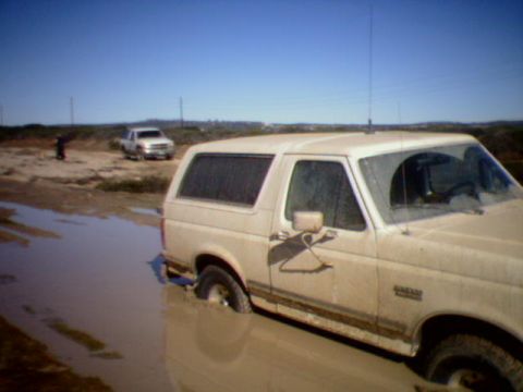 1990 Ford Bronco - A tan Ford Bronco is stuck in a muddy puddle with another white SUV in the background under a clear blue sky.