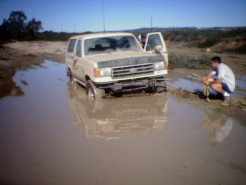 1990 Ford Bronco - A 1990 Ford Bronco is stuck in deep mud, with a person preparing to use a tow strap.