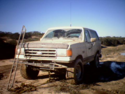 1990 Ford Bronco - This Ford Bronco is parked in a dusty, outdoor area with scrub brush and a body of water in the background.