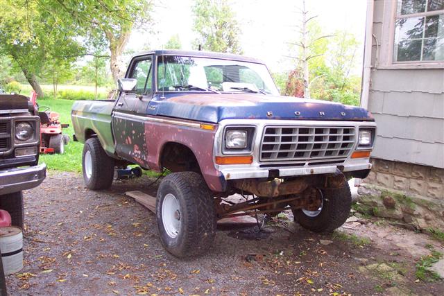 Lifted 1979 Ford F-100 pickup in mixed blue, red, and green primer panels on gravel driveway