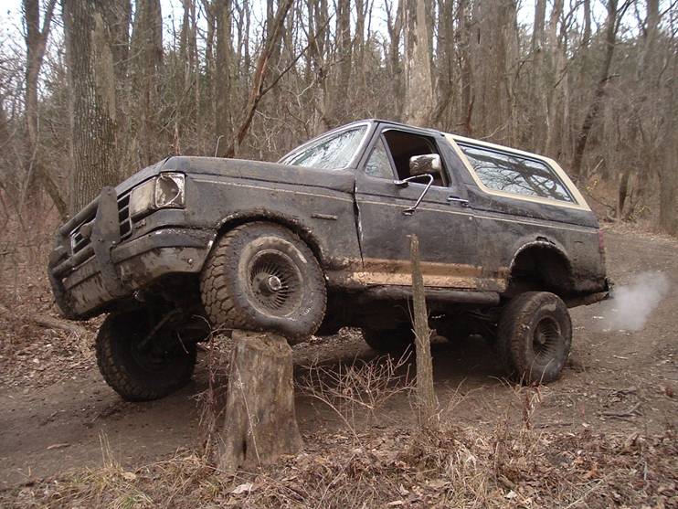 Muddy black lifted Ford Bronco with front tire on a tree stump in the woods