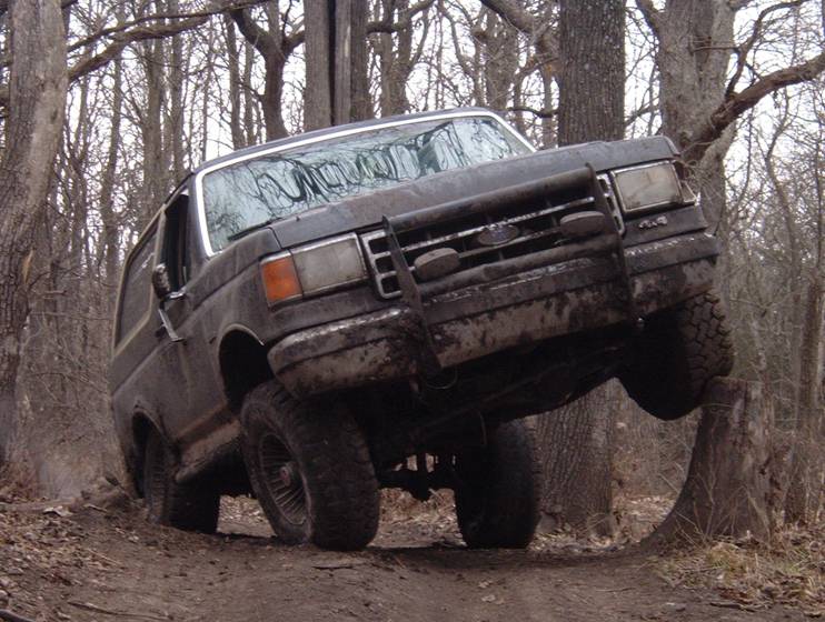 Lifted dark Ford Bronco with brush guard flexing on a muddy forest trail, front wheel in the air