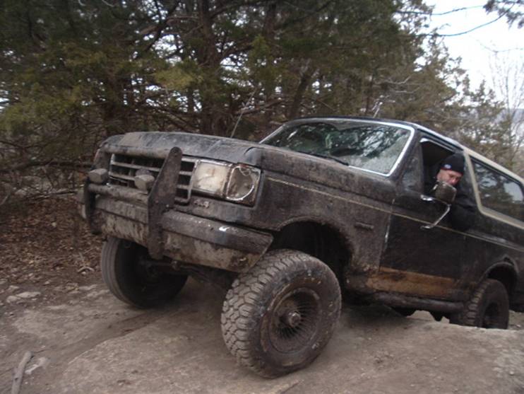 Mud-covered black Ford Bronco with brush guard crawling over rock ledge on a trail, driver visible