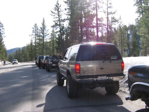 1985 Ford Bronco - Several lifted SUVs are parked in a line on a paved road with trees and mountains in the background.