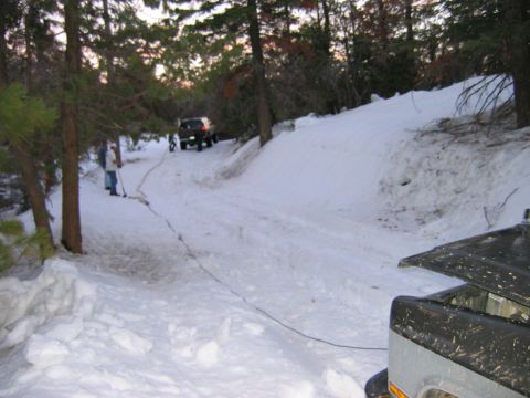 1985 Ford Bronco - Two SUVs are stuck in the snow on a wooded trail, with people attempting to free one with a tow rope.