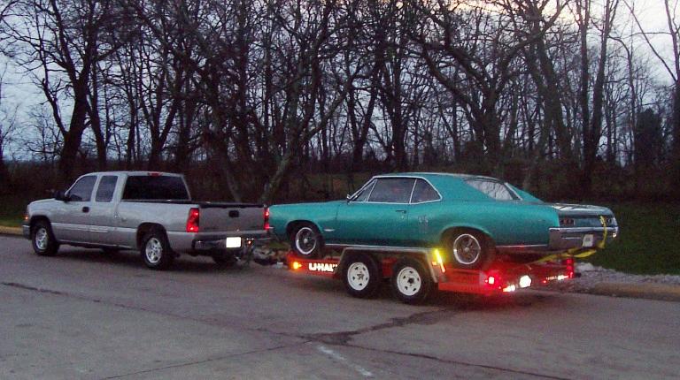 Teal coupe on red U-Haul trailer towed by silver pickup at dusk
