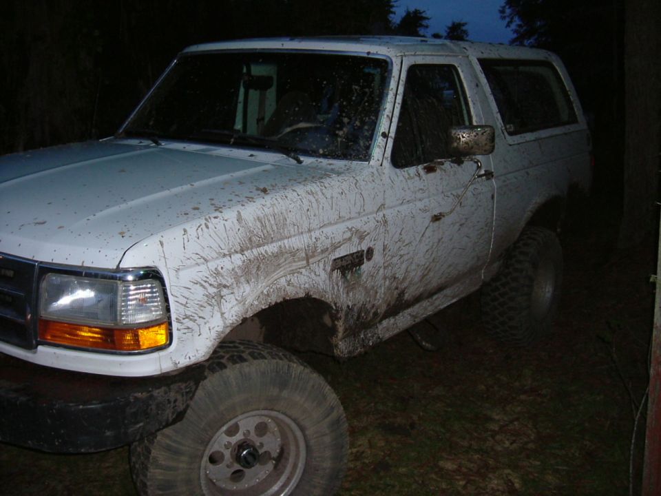 Mud-caked white 1990s Ford SUV on lifted suspension and mud tires at dusk