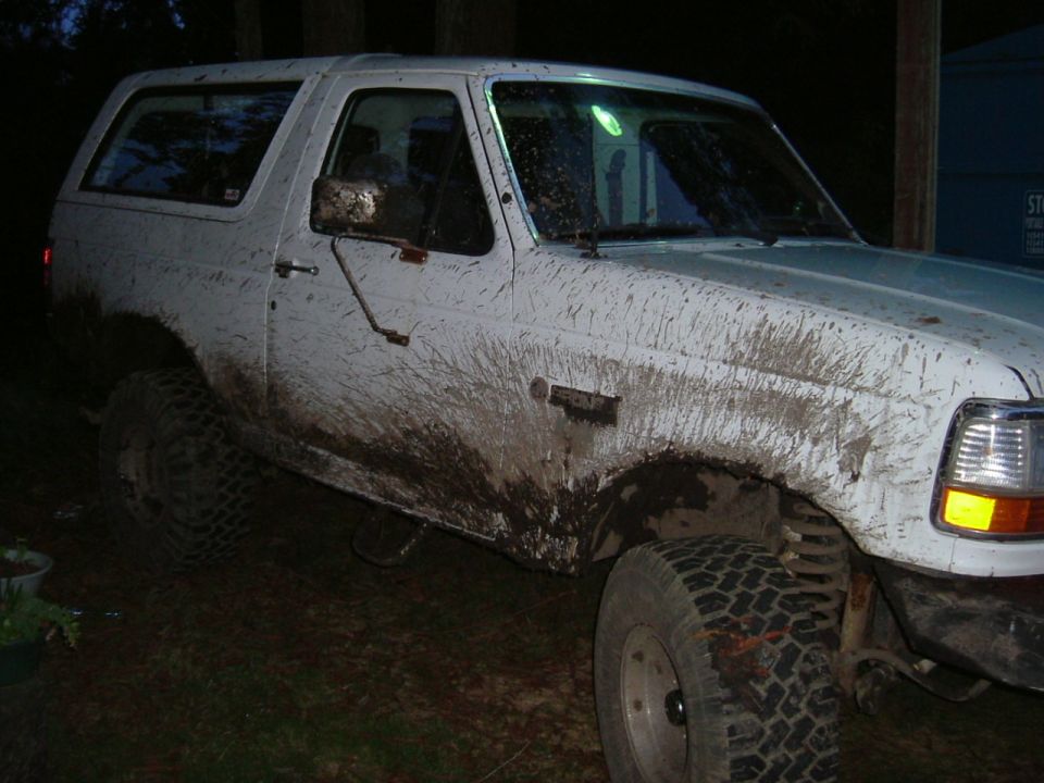 Mud-covered white Ford Bronco on lifted suspension and mud tires at night