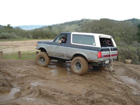 1989 Ford Bronco - A lifted Ford Bronco navigates a challenging muddy terrain with large off-road tires.