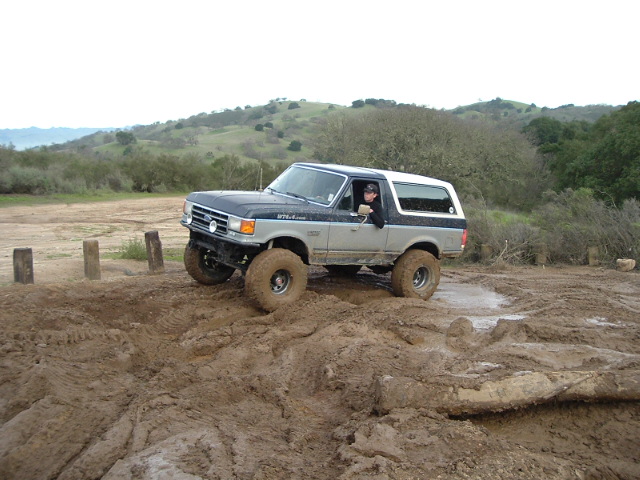 Black and silver lifted Ford Bronco on mud-terrain tires driving through deep mud off-road.