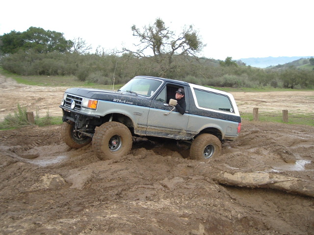 Gray and blue lifted Ford Bronco with white top driving through deep mud off-road