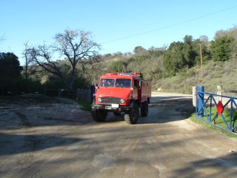 1989 Ford Bronco - A red Mercedes-Benz Unimog truck is seen driving on a dirt road with trees and hills in the background.