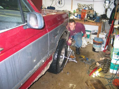 1995 Ford Bronco - Man working on the front tire of a 1995 Ford Bronco in a garage, surrounded by tools and car parts.