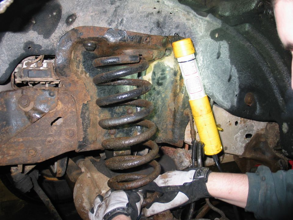 Close-up of rusted suspension coil and bracket, illuminated by a yellow flashlight with visible text