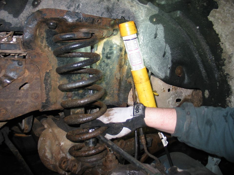Close-up of a heavily rusted vehicle suspension system, showing a coil spring and a yellow shock absorber. A gloved hand is visible near the spring