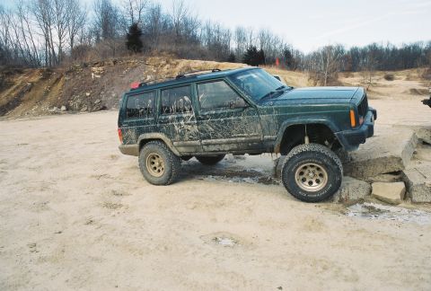 1995 Ford Bronco - A muddy Jeep Cherokee XJ navigates an obstacle course in a dirt lot.